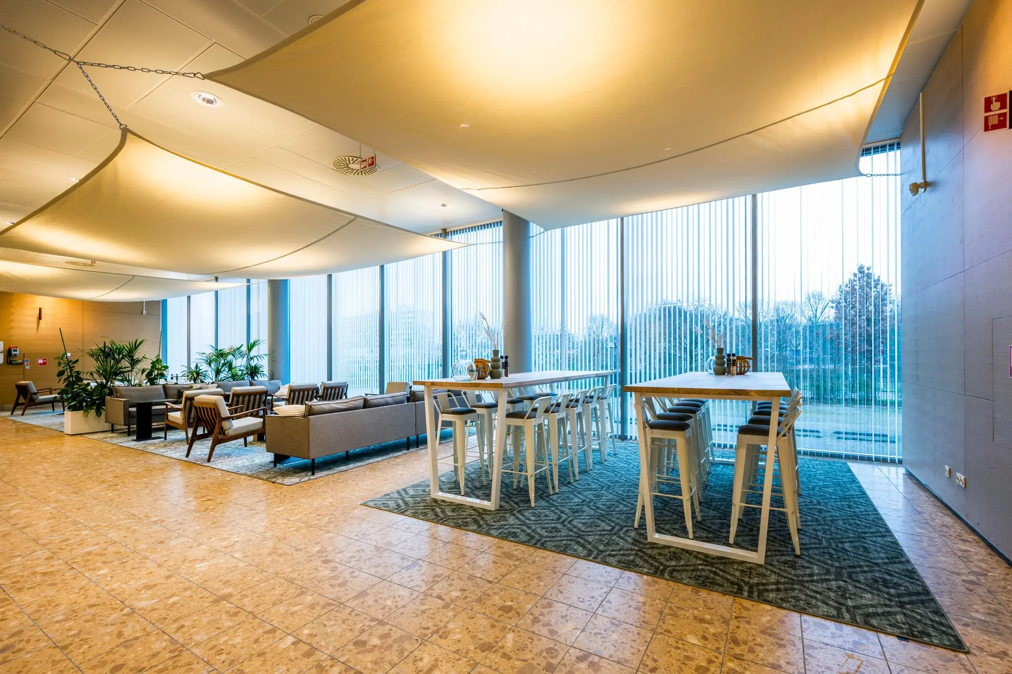 Moderne wachtruimte of ontmoetingsruimte aan de Burgemeester Rijnderslaan met zithoeken, hoge tafels en grote ramen met verticale jaloezieën.